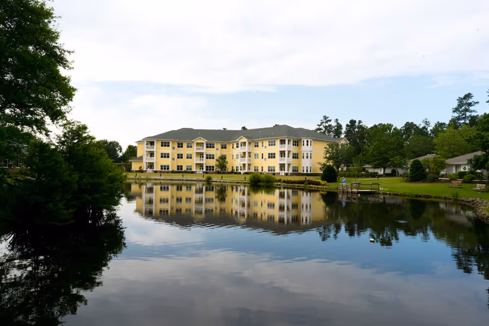 Yellow multi-story senior living building sits beside a calm lake with trees and reflections.