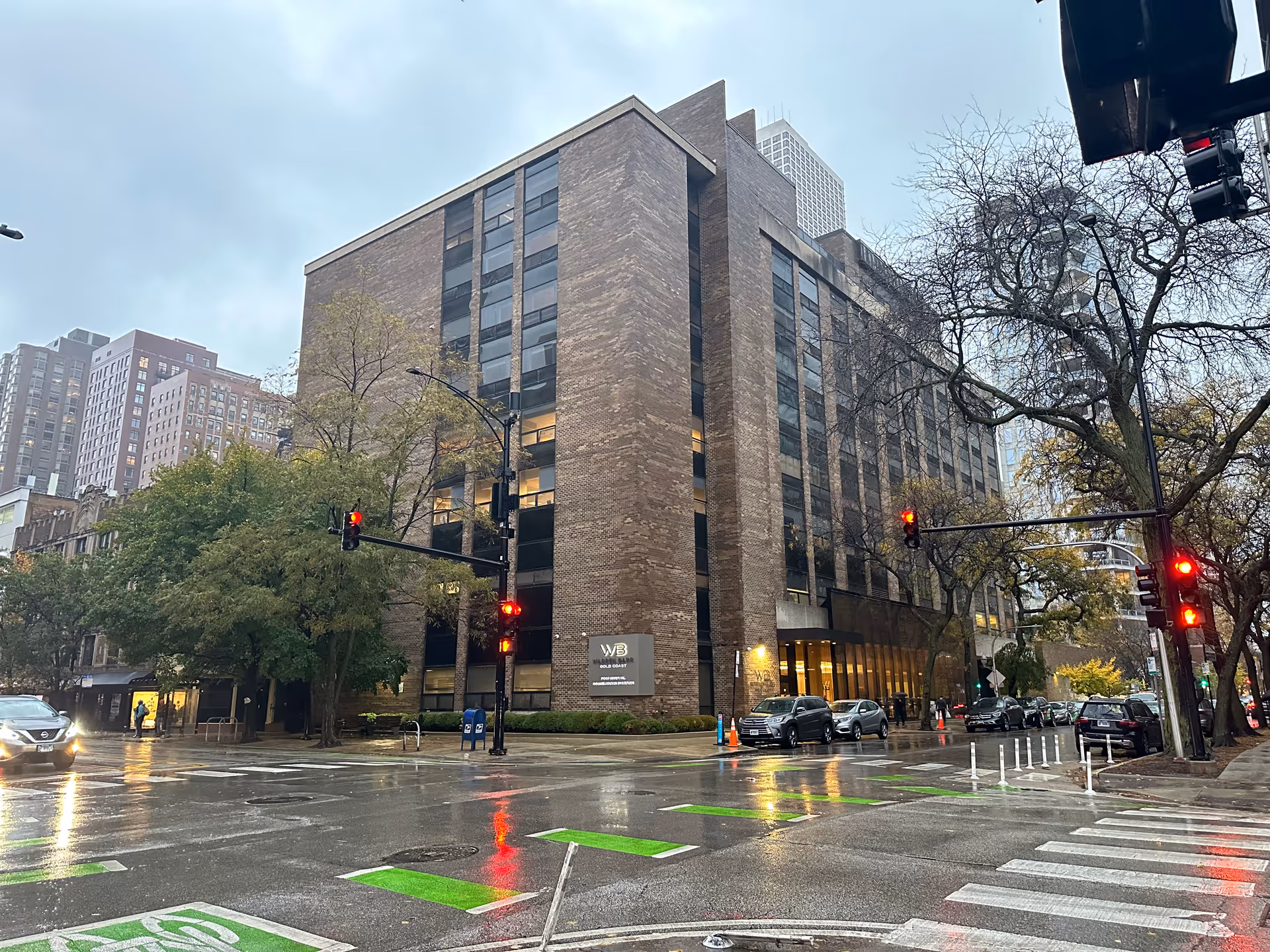 Rainy street corner showing a multi-story brick apartment building with an entrance, cars, traffic lights, and wet crosswalks.