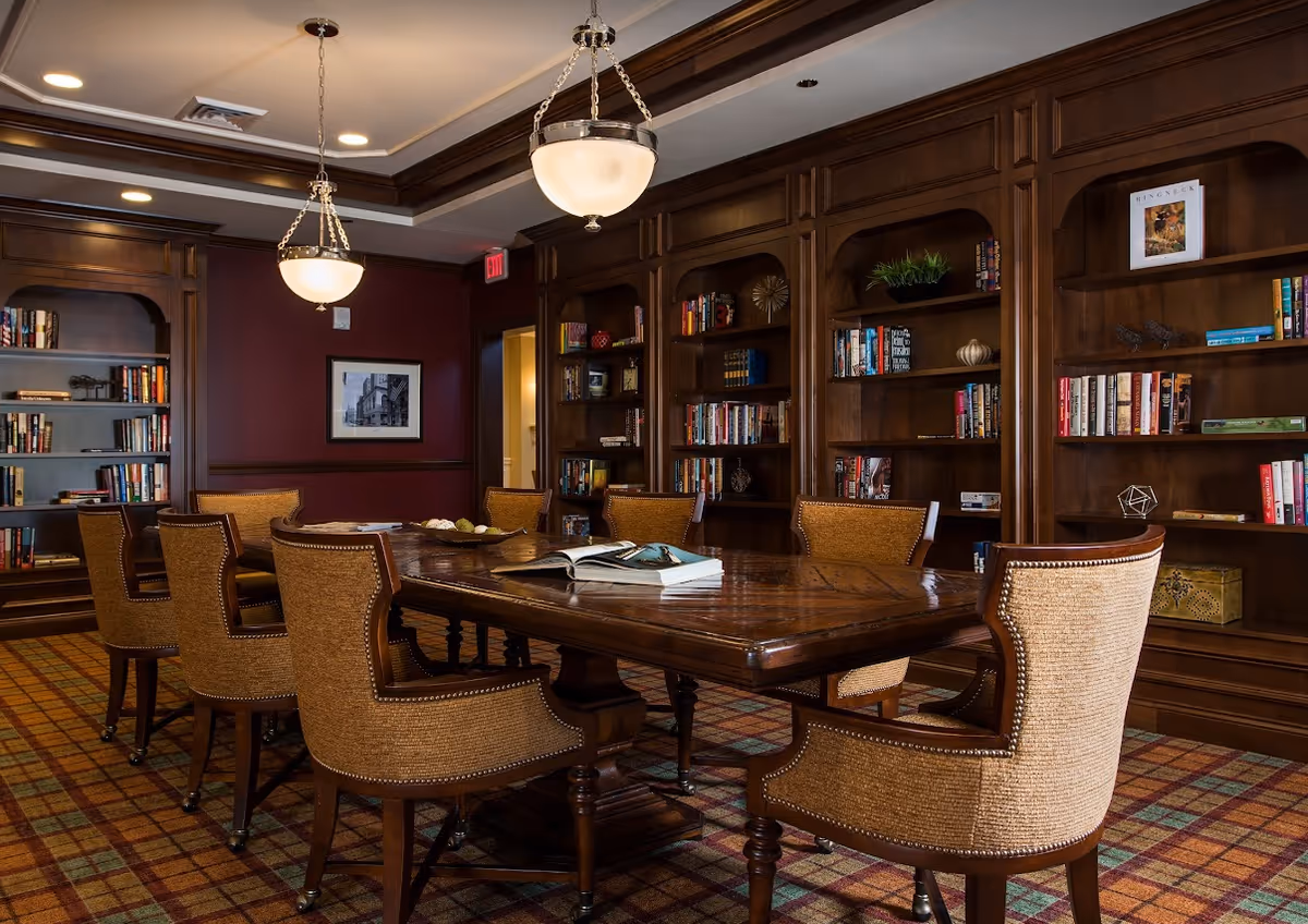 A cozy library or meeting room with a large wooden table surrounded by eight upholstered chairs. The walls are lined with built-in wooden bookshelves filled with books and decorative items. Two pendant lights hang from the ceiling, and the room has a warm, inviting atmosphere with a plaid carpet and dark wood paneling.