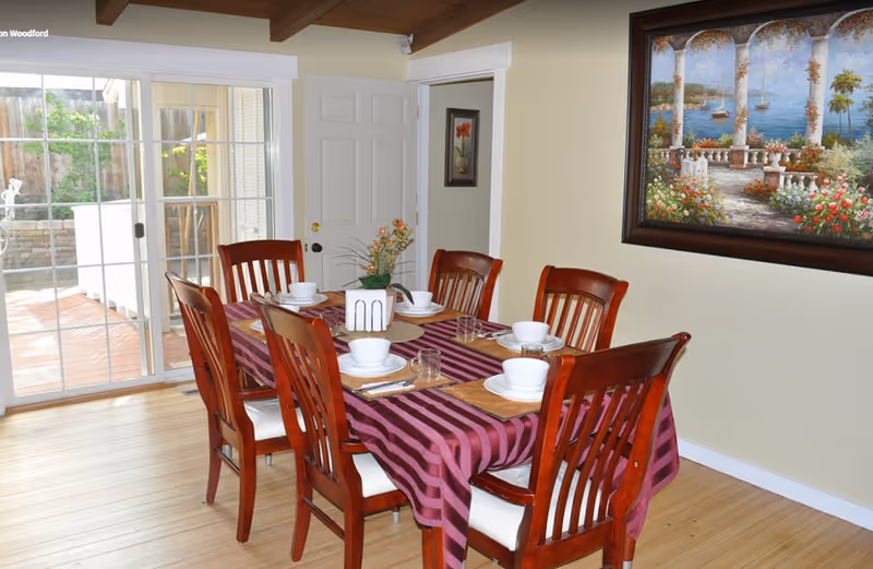 Dining room with a wooden table set for six covered by a purple striped tablecloth, sliding glass doors, and a large framed painting on the wall.