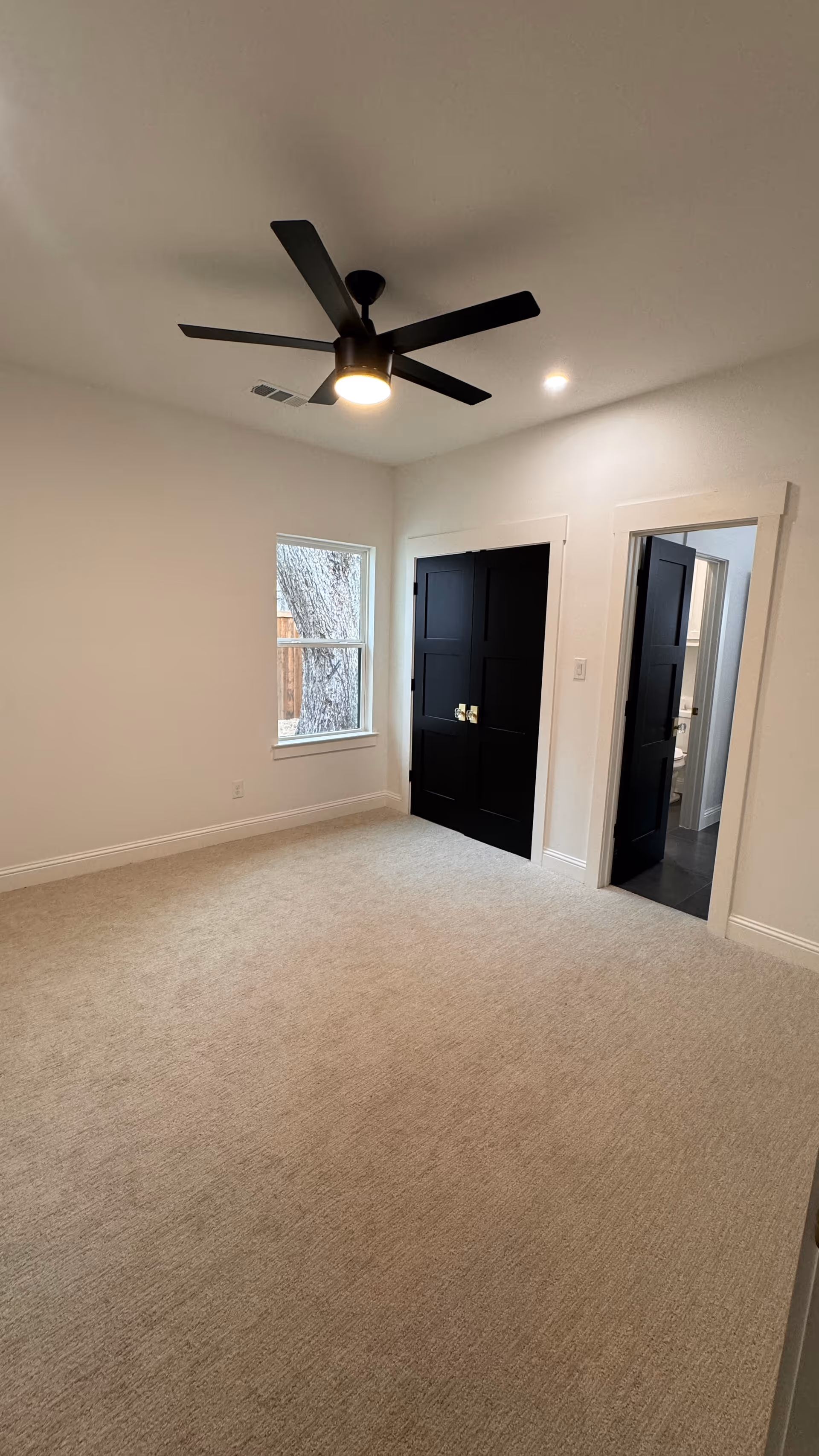 Empty room with beige carpet, white walls, a window showing a tree outside, black double doors, an open doorway leading to a bathroom, and a modern black ceiling fan with light.