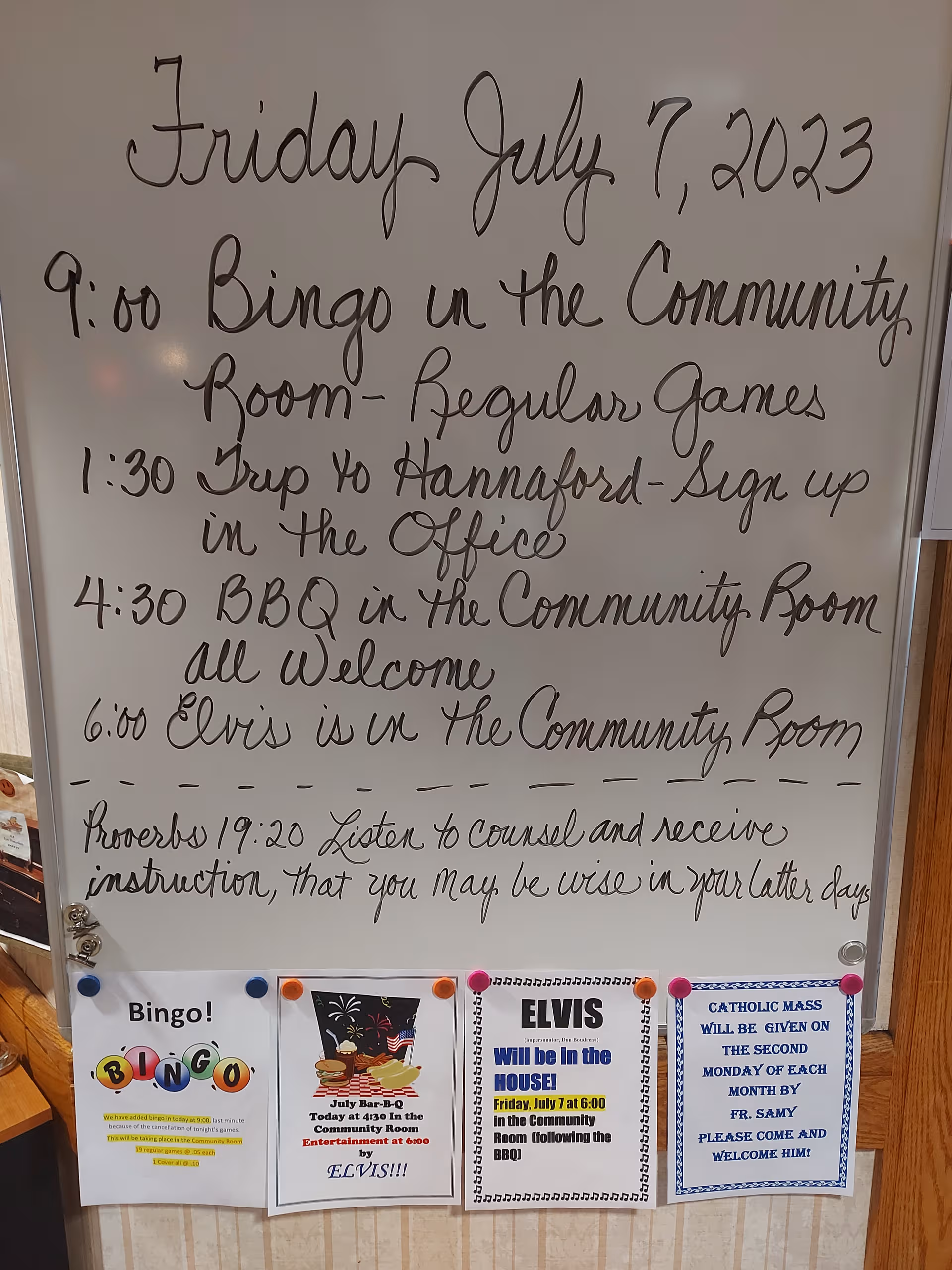 Whiteboard in Montello Heights lobby shows Friday schedule and posters for bingo, Elvis, and BBQ.