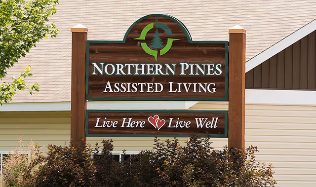 Wooden sign for Northern Pines Assisted Living with a green tree logo above the text. The sign reads 'NORTHERN PINES ASSISTED LIVING' and below it says 'Live Here ♥ Live Well'. The sign is mounted on two wooden posts with a building and some bushes in the background.