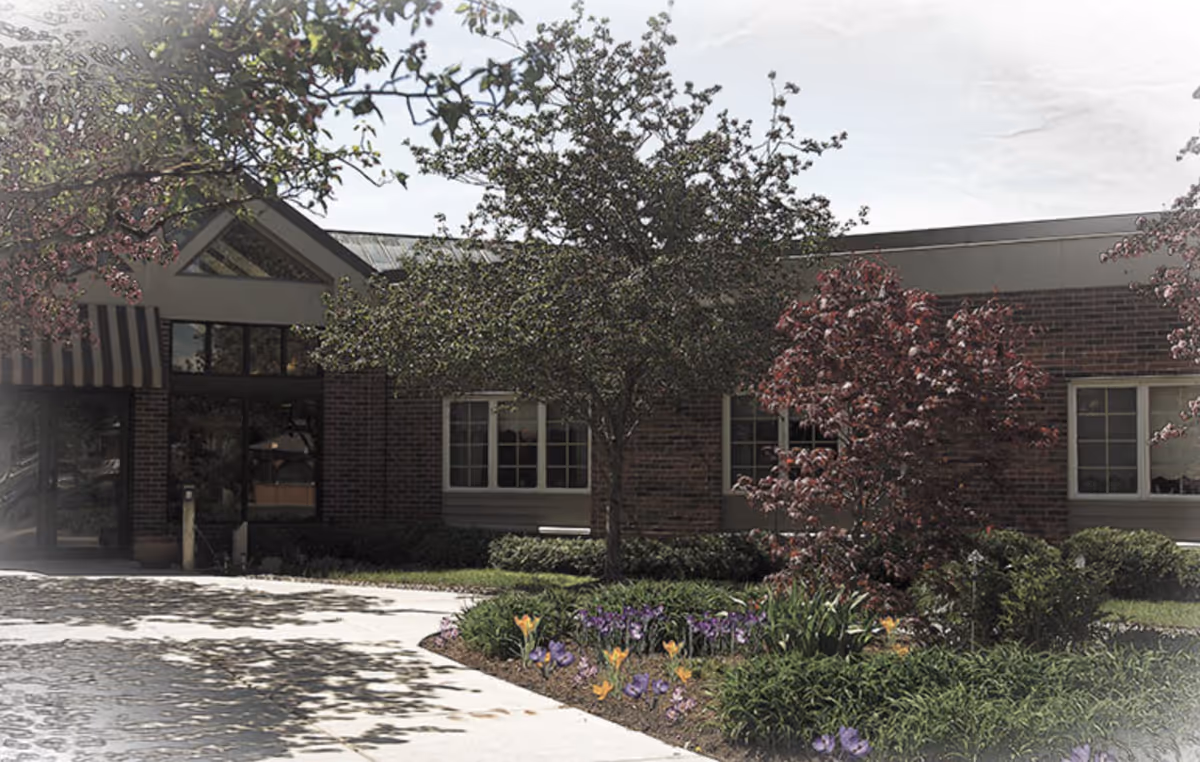 Exterior front entrance of a single-story brick senior living facility with trees, flowering beds, and a sidewalk.