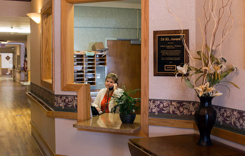 Reception area inside Northcrest Rehab and Nursing Center with a woman sitting behind the counter talking on the phone. The area features wooden trim, a potted plant on the counter, a large vase with flowers on a nearby table, and a plaque on the wall. A hallway extends into the background.