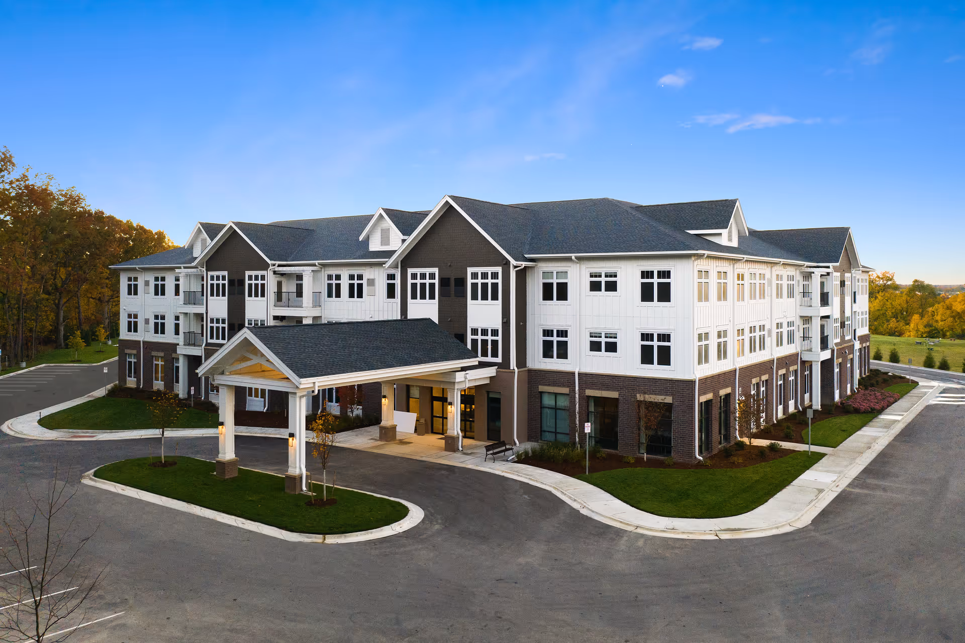 Exterior view of a large, modern three-story senior living facility building with a covered entrance, surrounded by paved driveways and landscaped greenery under a clear blue sky.