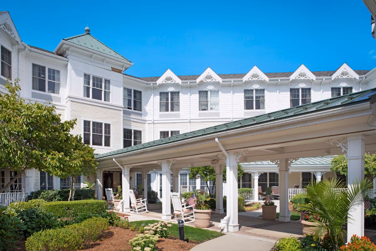 White three-story senior living building with a covered porte-cochère, rocking chairs, and a landscaped courtyard under a clear blue sky.