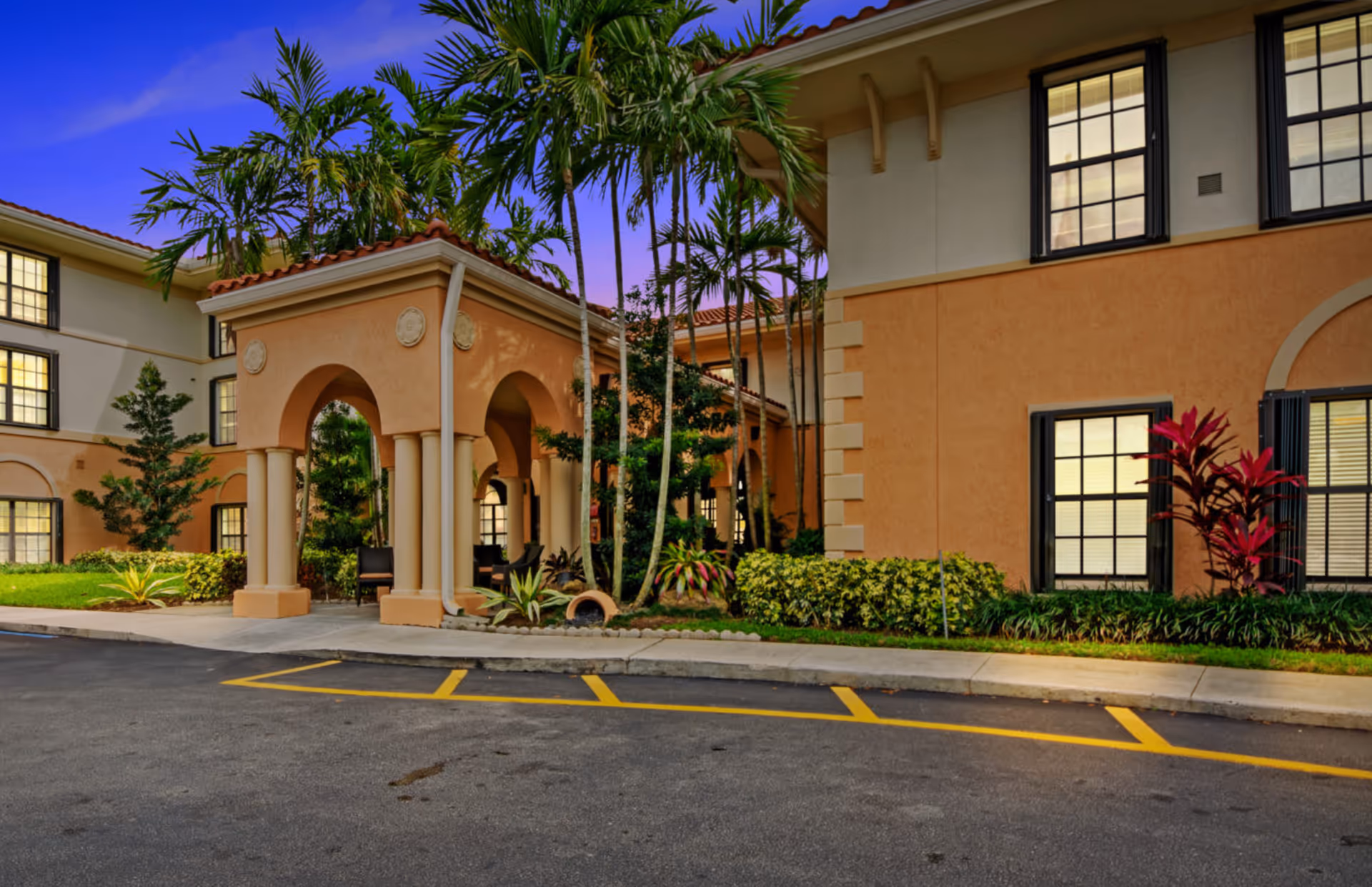 Front exterior of a Mediterranean-style senior living building with an arched covered entrance, palm trees, and a parking area at dusk.