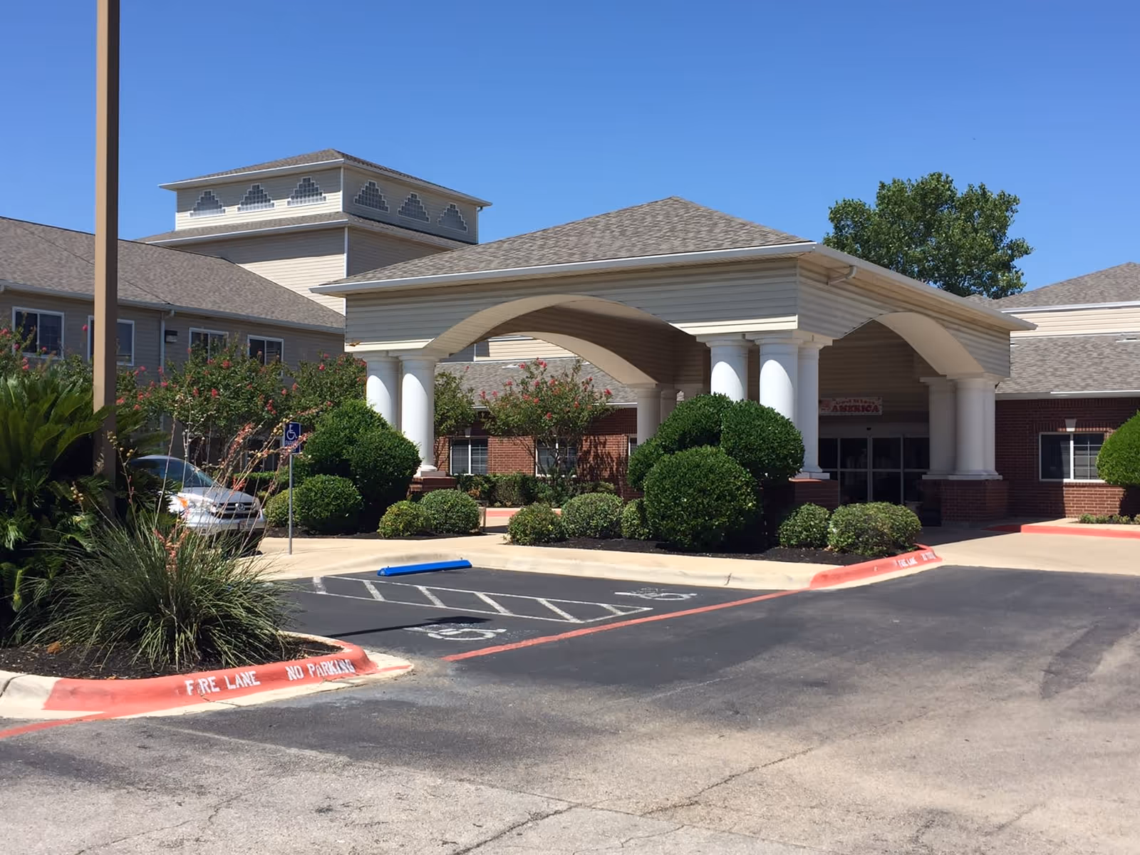 Exterior view of the entrance to Garden Estates of Temple, showing a covered drop-off area with white columns, manicured bushes, and a clear blue sky.