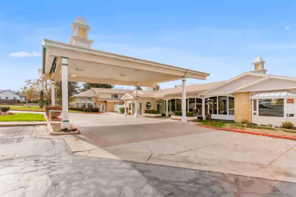 Exterior view of The Beaumont Assisted Living and Memory Care facility showing the entrance with a large covered drop-off area, beige building with white trim, and clear blue sky.