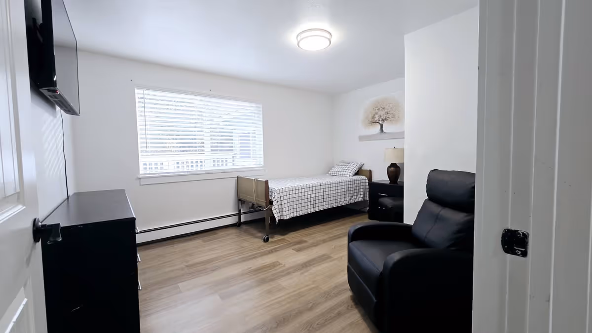 Sunlit senior living bedroom with a bed, recliner, dresser, and wall-mounted TV.