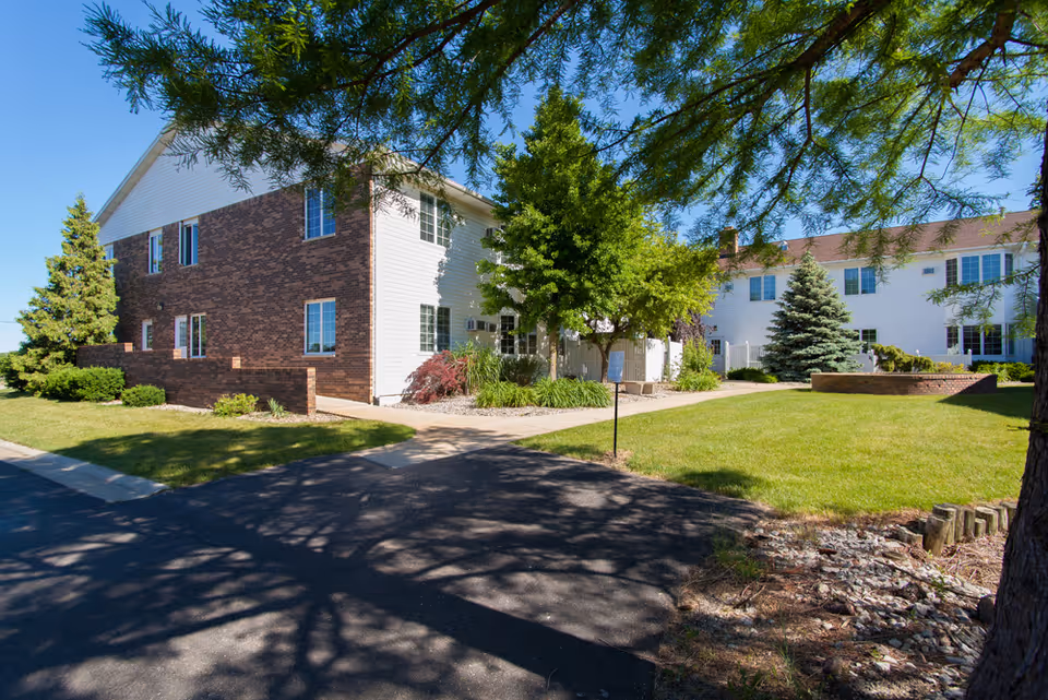 Exterior view of a two-story brick and white-siding senior living building with a landscaped courtyard, trees, and a paved driveway.