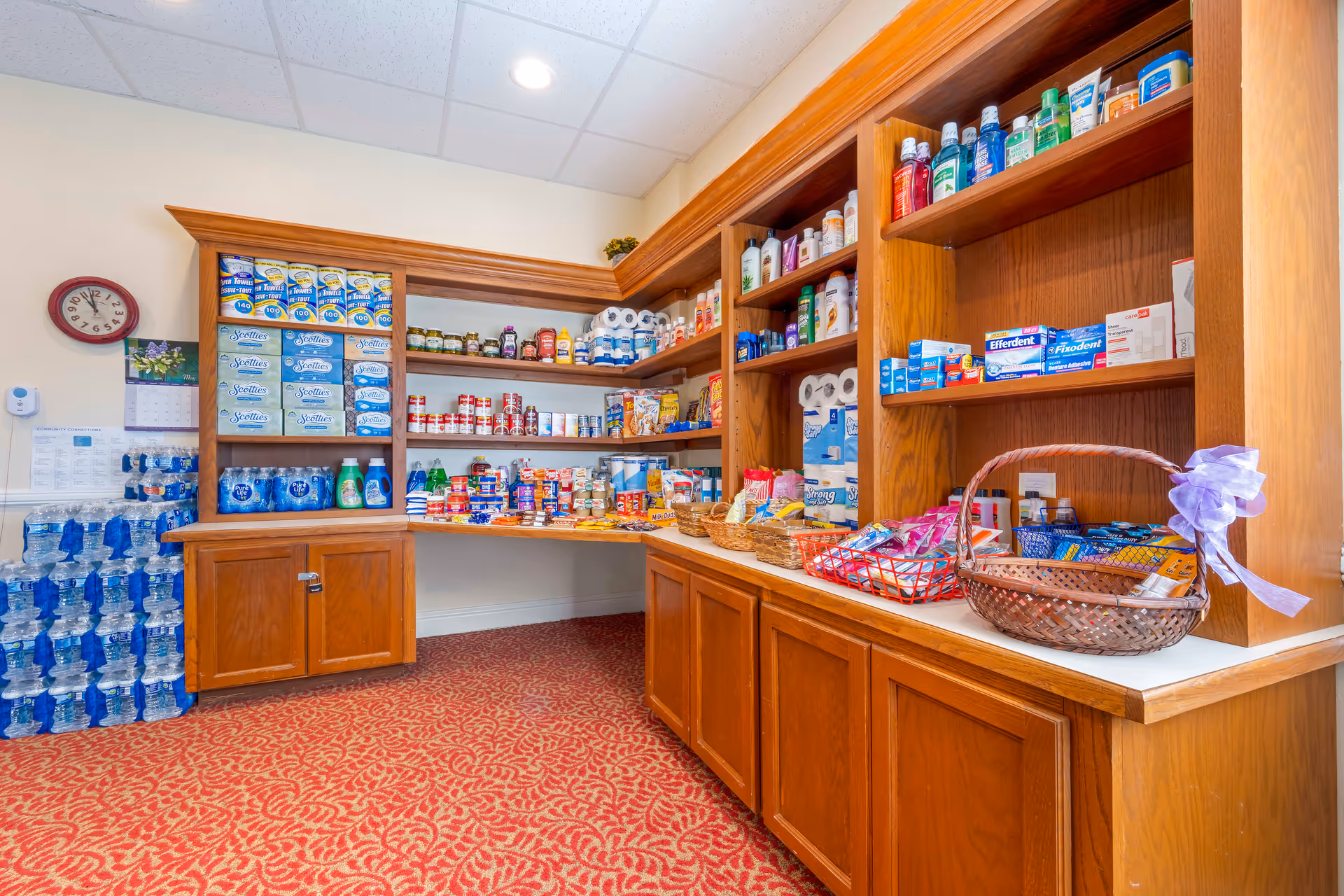 A well-stocked pantry area with wooden shelves and cabinets containing various food items, bottled water, paper towels, and personal care products. The room has a red patterned carpet and a clock on the wall showing the time as 9:10.