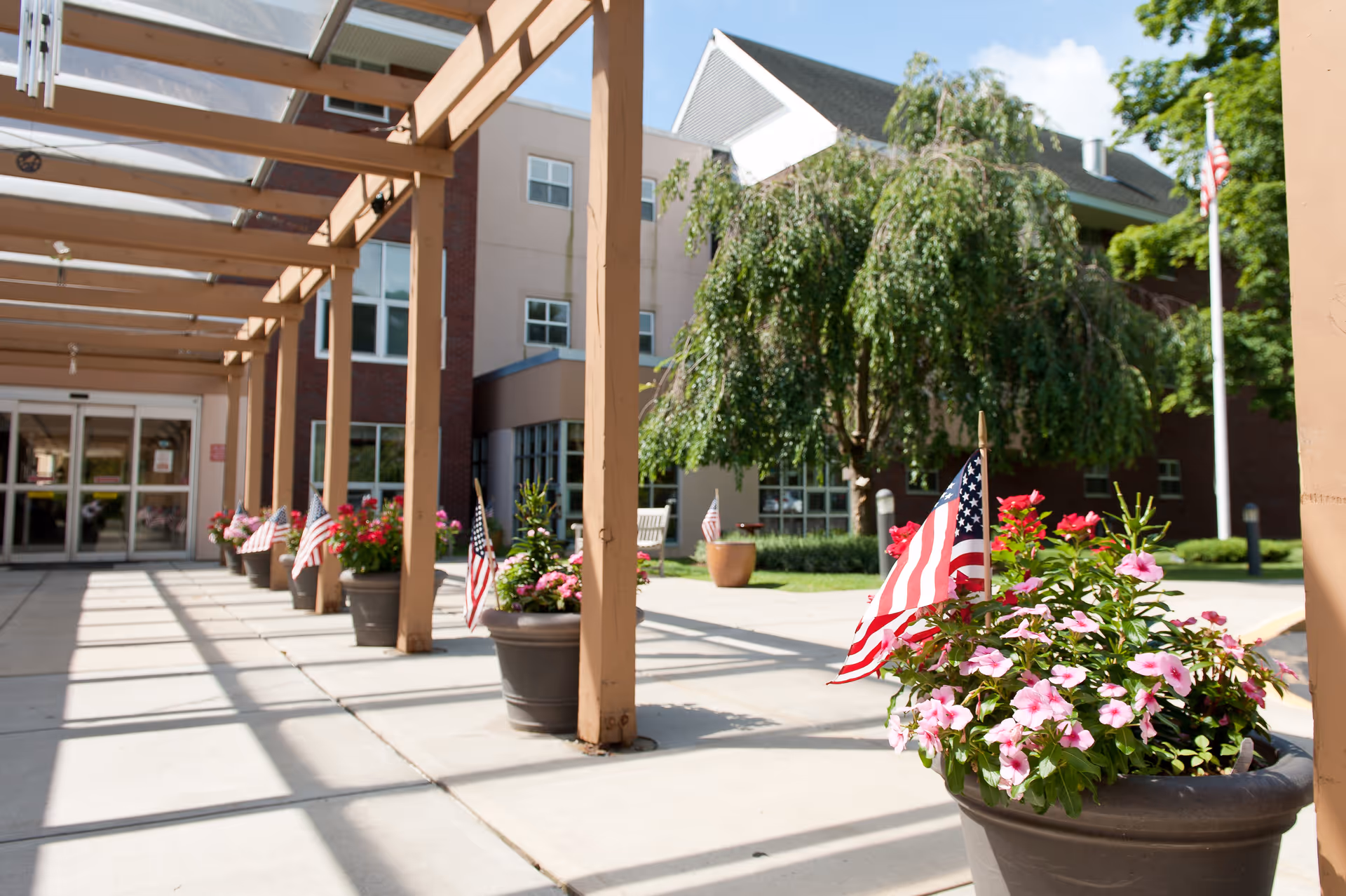 Covered entrance walkway with potted flowering plants and small American flags leading to the front of a multi-story building.