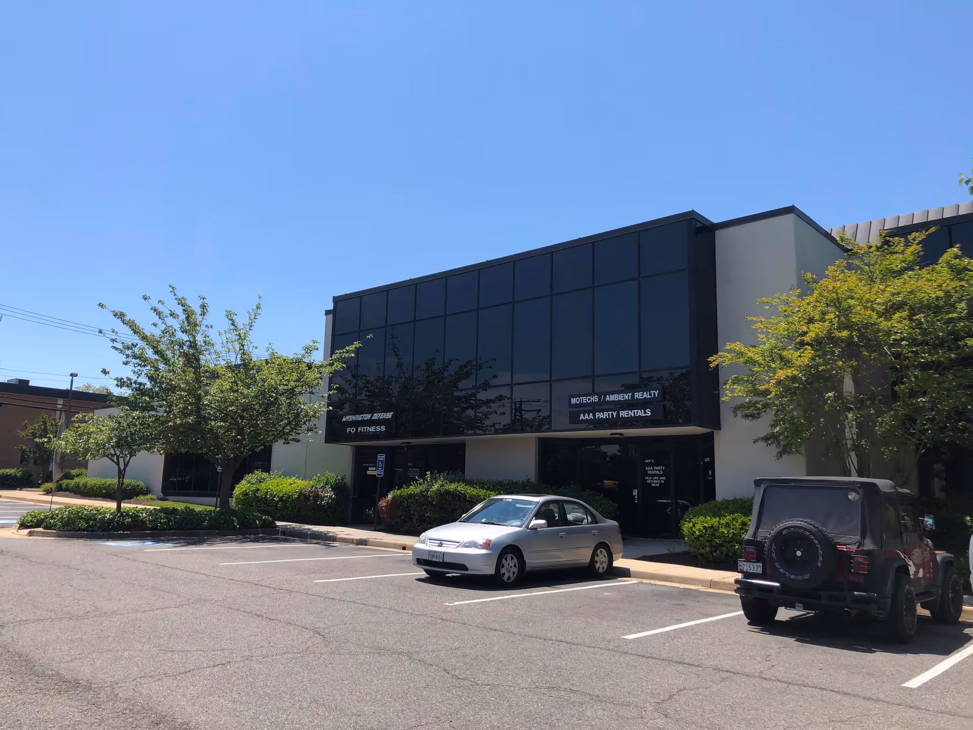 Modern glass-front building with parking lot and trees on a sunny day