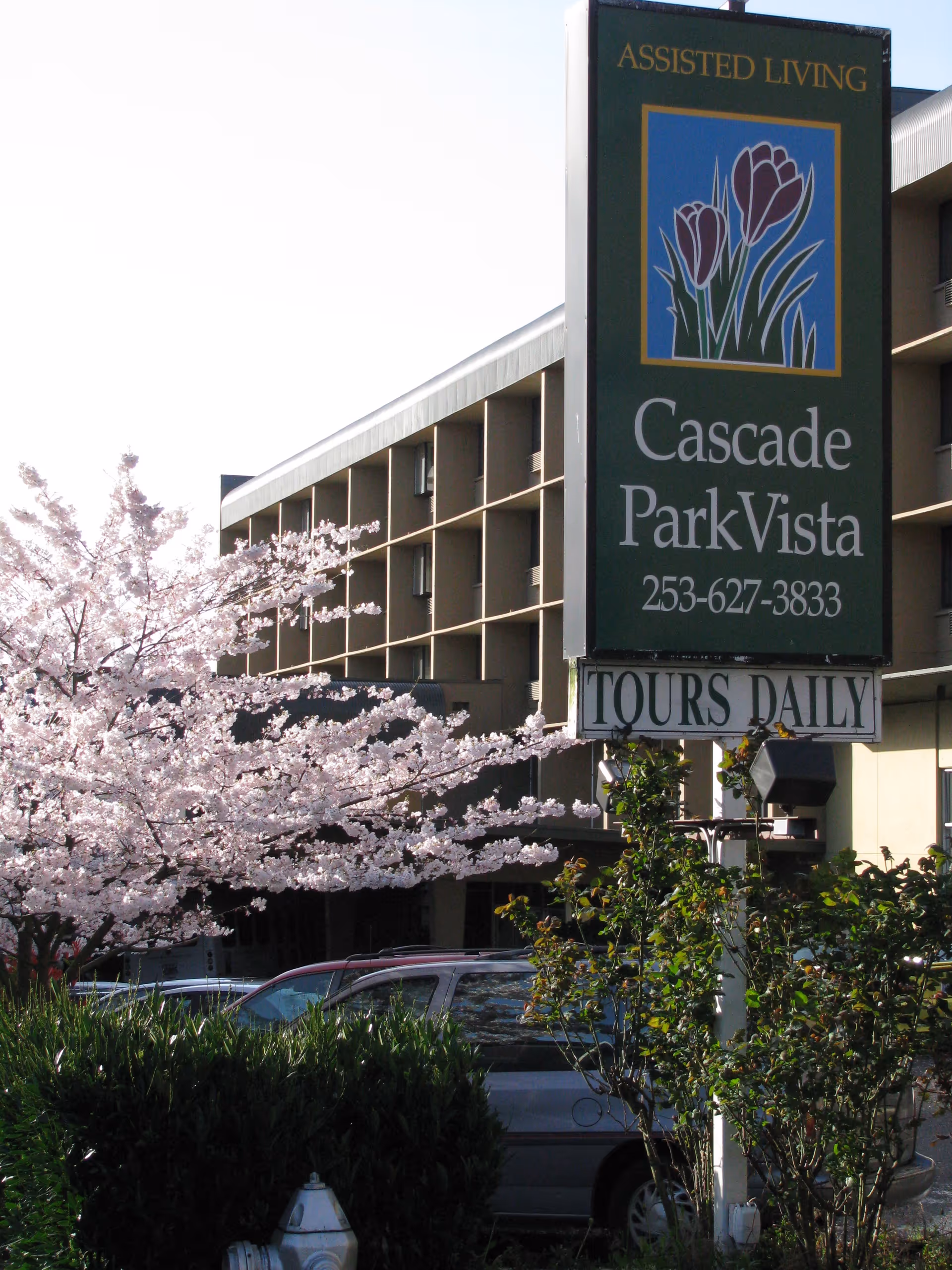 Exterior view of Cascade Park Vista assisted living facility with a large sign displaying the facility name, phone number, and 'Tours Daily'. There is a blooming tree with pink flowers and some parked cars in front of the building.