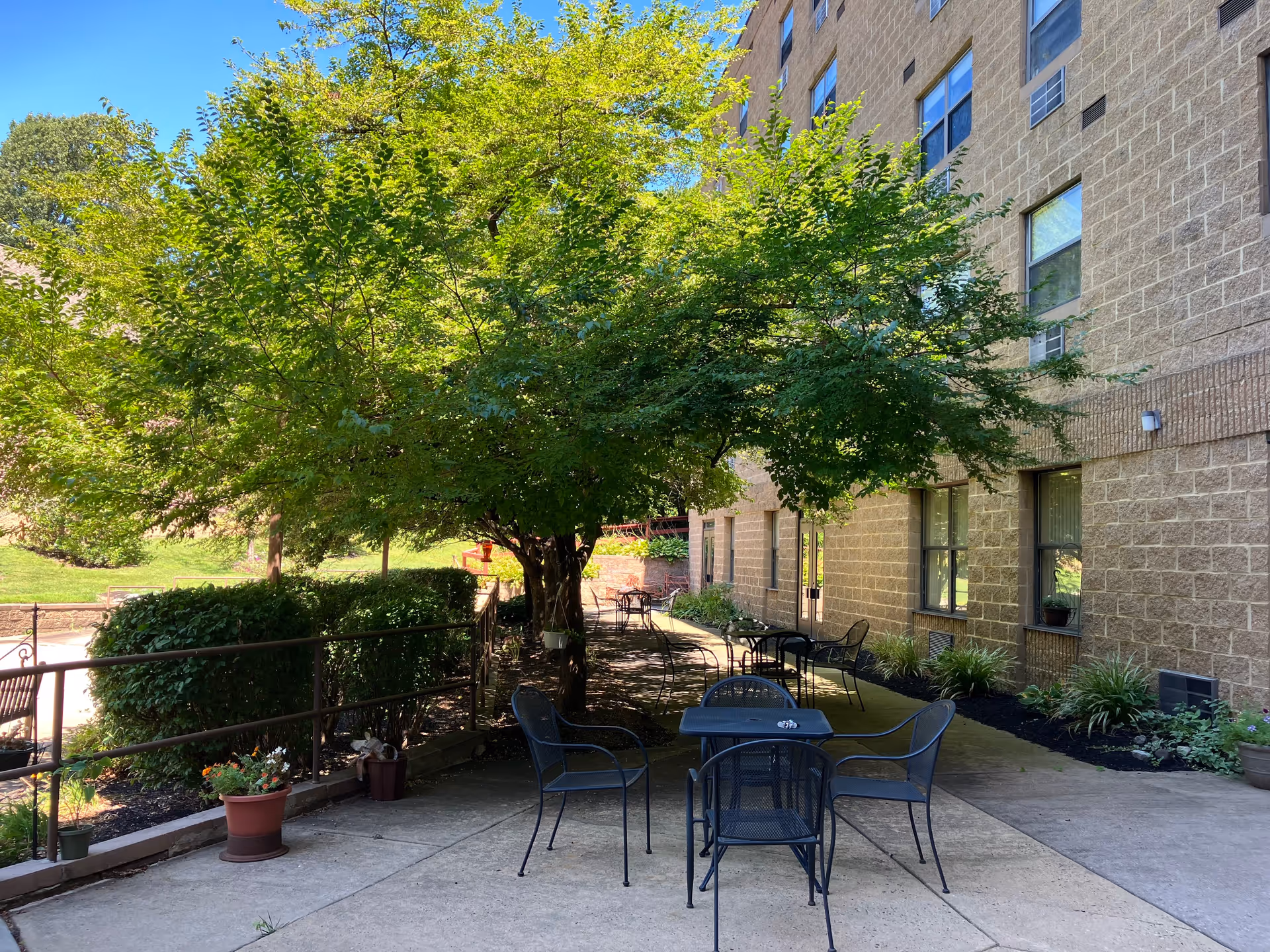 Outdoor patio area at Gloria Dei Manor with metal tables and chairs arranged on a concrete surface. There are large leafy trees providing shade, potted plants along the edge, and a beige brick building with windows in the background.