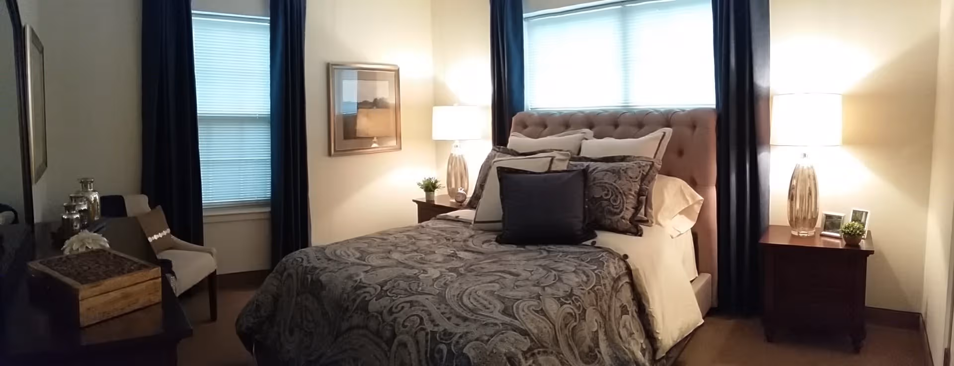 Bedroom with tufted headboard, patterned bedding, two nightstands, lamps, and dark curtains.