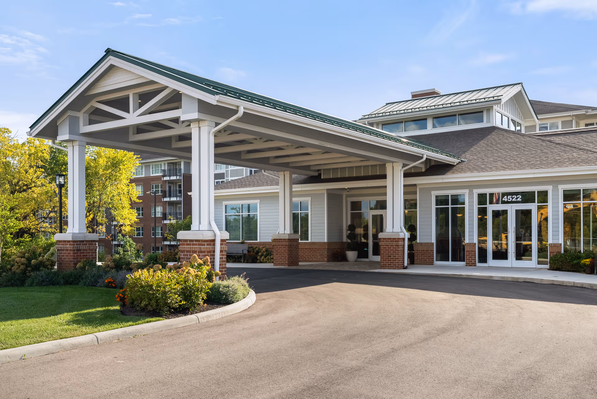 Entrance of a senior living facility with a covered drop-off area supported by white columns with brick bases. The building has large windows and a door with the number 4522 above it. There are landscaped bushes and trees around the driveway under a clear blue sky.