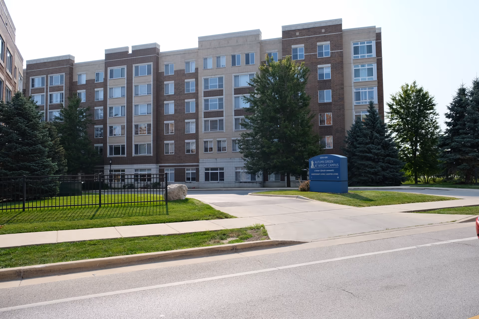 Front exterior of a multi-story brick senior living building with trees, a lawn, driveway, and a blue entrance sign.