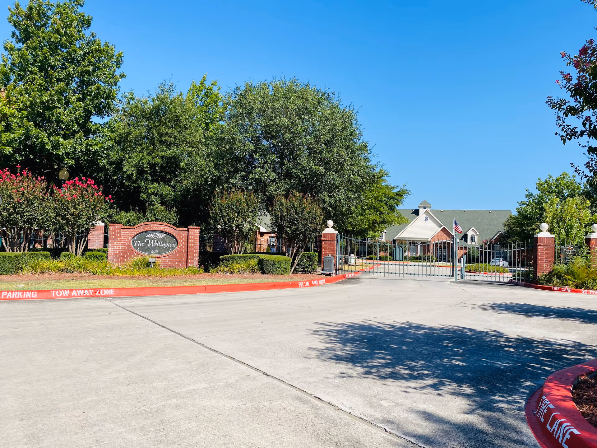 Entrance to The Wellington senior living facility featuring a gated driveway, brick pillars, manicured bushes, trees, and a clear blue sky.