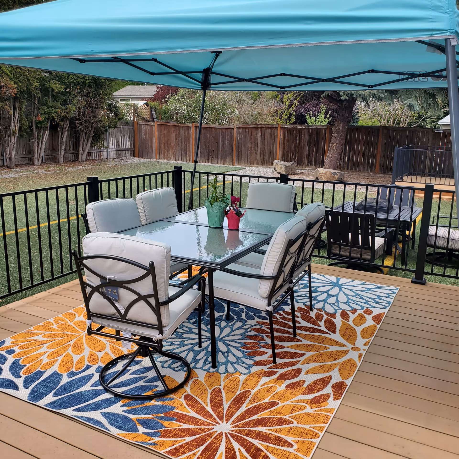 Outdoor patio with glass table, cushioned chairs, colorful rug, under teal canopy
