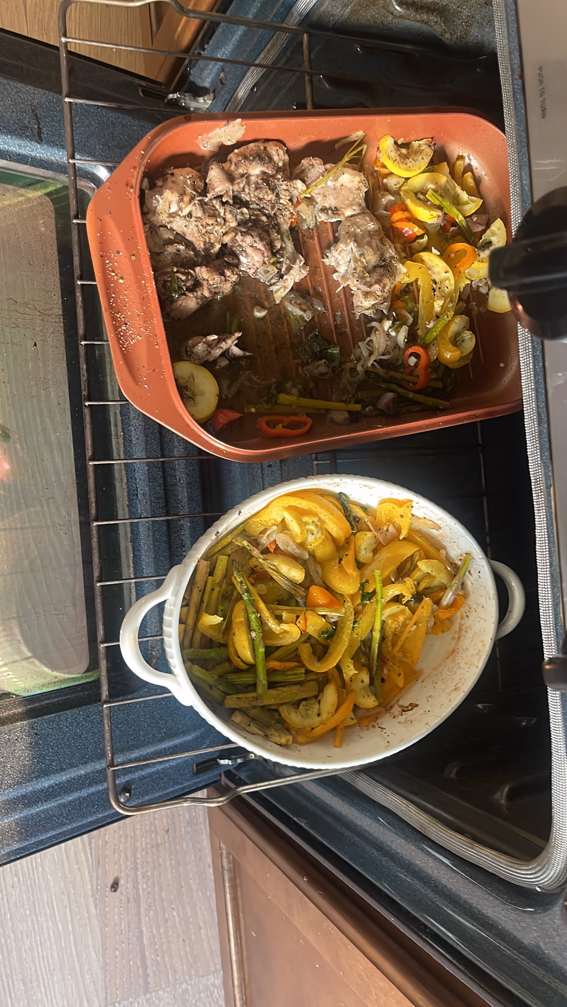 Two roasting dishes with cooked meat and yellow squash and asparagus inside an open oven.