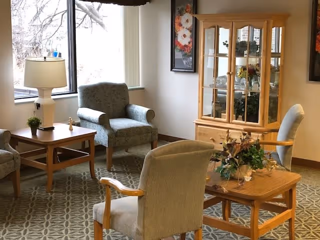 A cozy sitting area with four upholstered armchairs arranged around two wooden tables. One table has a lamp and a small plant, and the other has a floral arrangement. Behind the chairs is a wooden display cabinet with glass doors containing decorative items. The room has large windows letting in natural light and framed floral artwork on the walls.