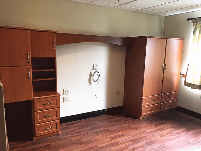 Empty room with wooden floor and built-in wooden cabinets and drawers along the walls, a window with curtains on the right side letting in natural light, and electrical outlets on the wall.