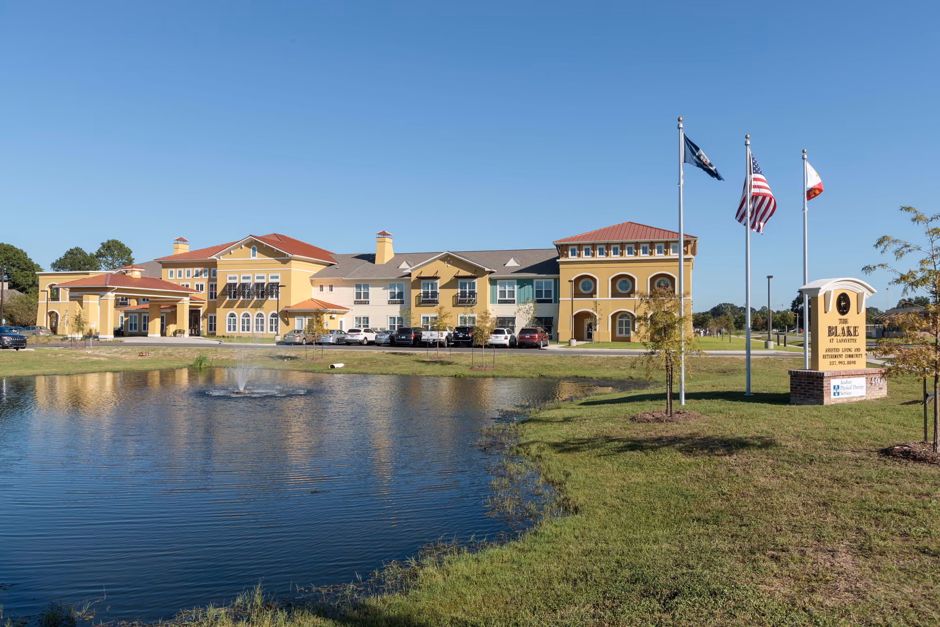 Front exterior of The Blake at Lafayette senior living building beside a pond, flags, and an entrance sign.