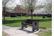 Outdoor park-like area with a bench along a sidewalk and trees near a building.
