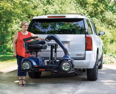 An older woman stands next to a mobility scooter attached to a white SUV outdoors.