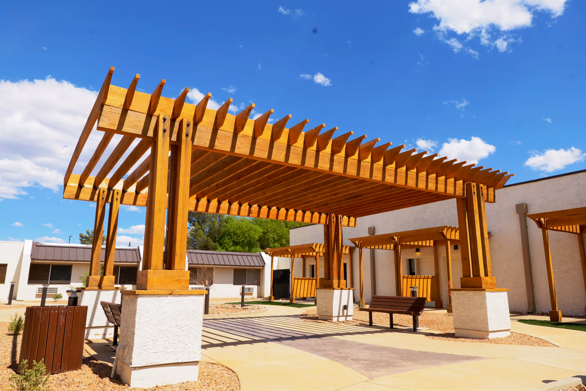 Outdoor courtyard area with wooden pergolas providing shade over benches and walkways, surrounded by single-story buildings under a blue sky with scattered clouds.