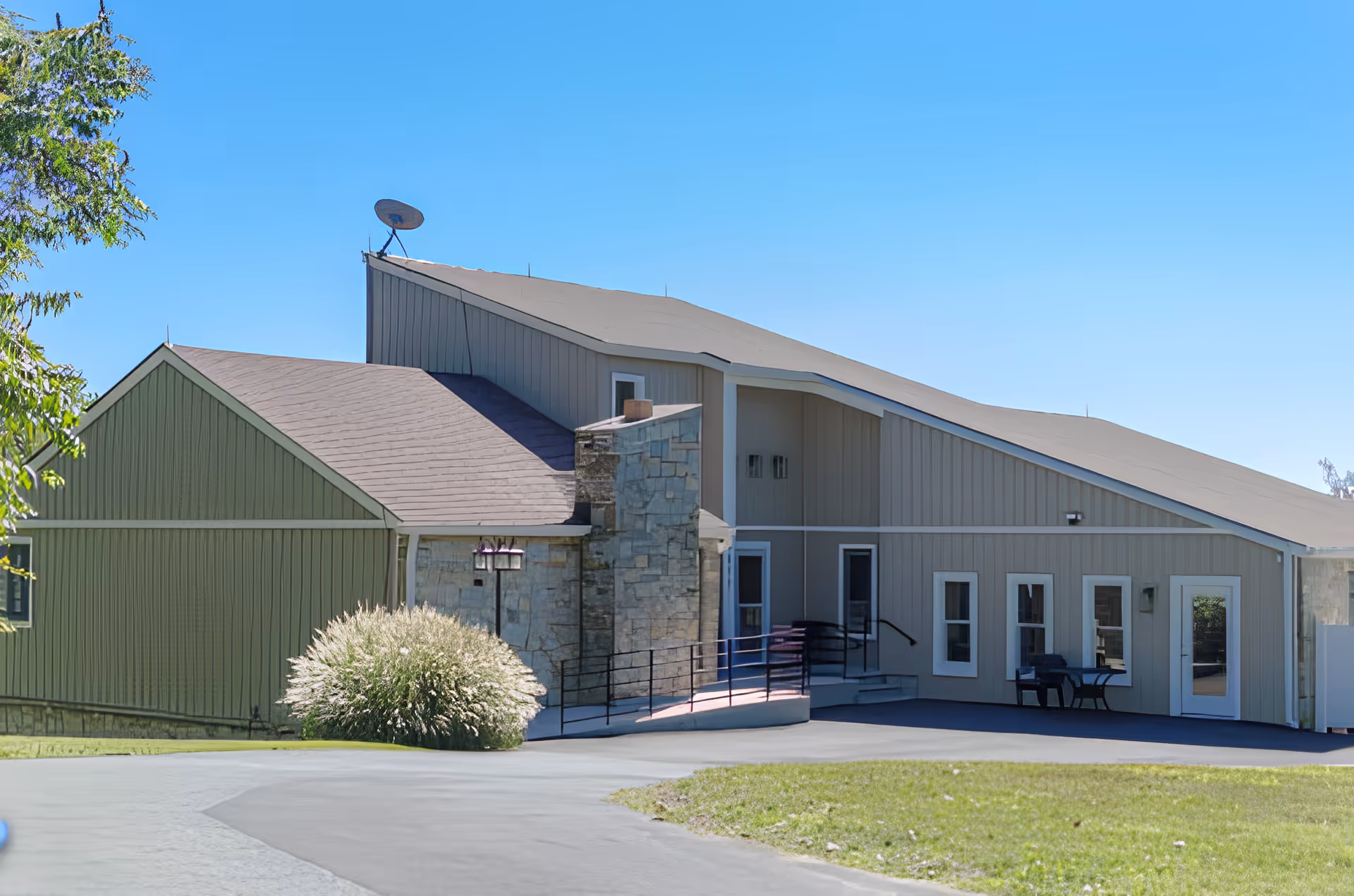Single-story assisted living building with sloped roofs, a stone entrance, wheelchair ramp and outdoor seating under a clear blue sky.