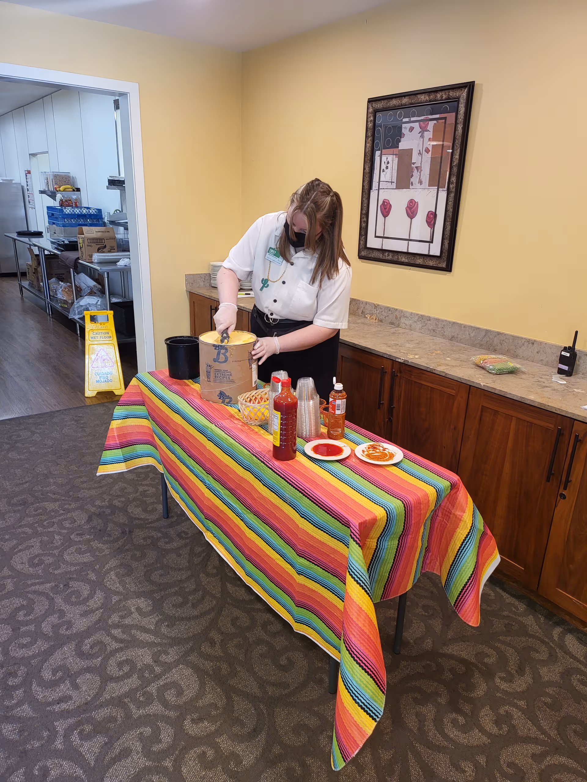 A staff member wearing a mask prepares food at a colorful table inside a senior facility.