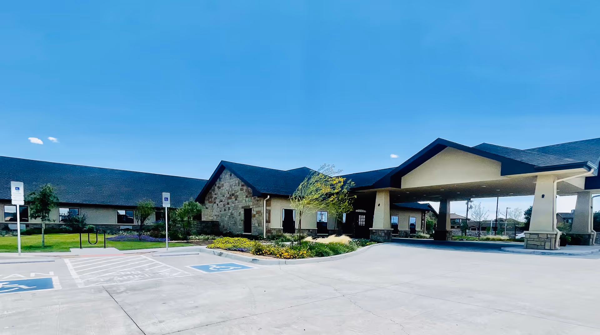 Exterior view of a single-story building with a covered entrance and a parking area in front, including handicap parking spaces. The building has a combination of stone and beige siding with a dark roof, surrounded by landscaped greenery and a clear blue sky above.