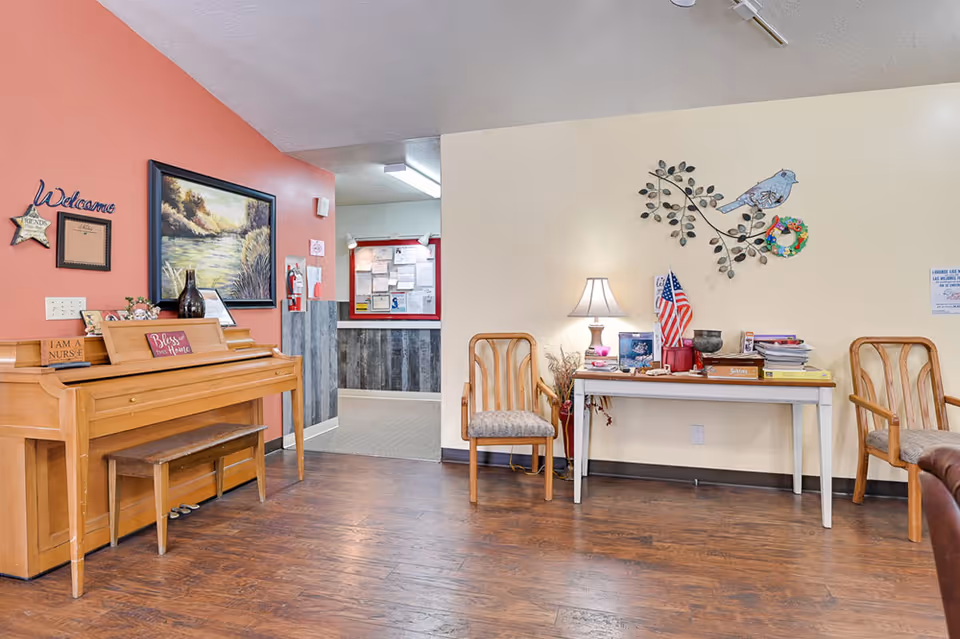 Interior view of a common area in an assisted living facility featuring a wooden piano with a bench, a painting above it, and a welcoming sign on the wall. There is a white table with various items including a lamp, an American flag, and books, flanked by two wooden chairs with cushioned seats. The floor is wooden, and the walls are painted in warm tones with decorative elements such as a metal bird wall art and a small wreath.