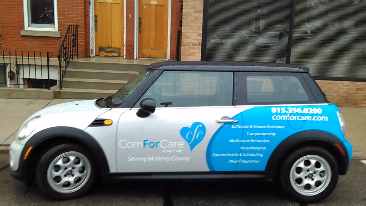 White and blue ComForCare home care vehicle parked on city street near brick building