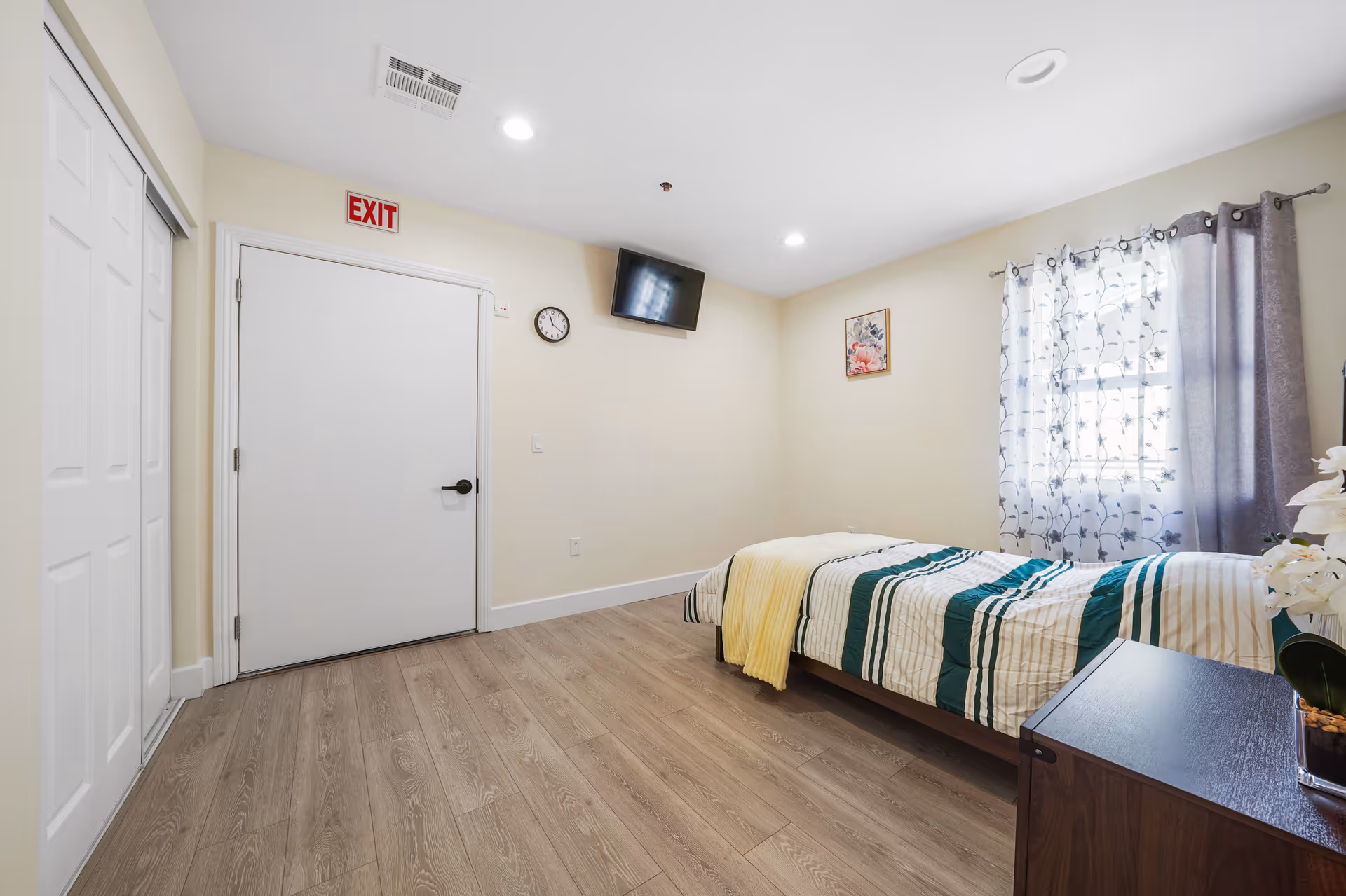 Bright, minimal bedroom with bed, dresser, window with patterned curtains, and exit door.