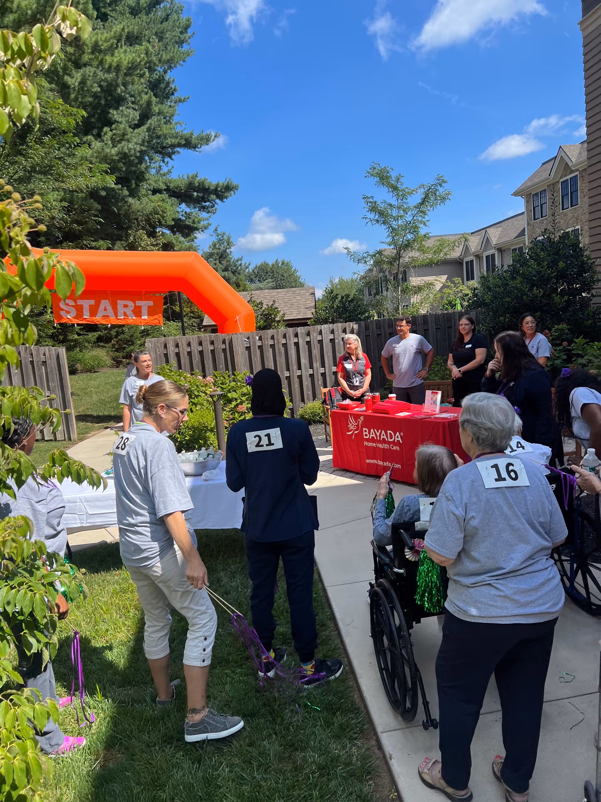 People gather outdoors around an orange start arch near a red BAYADA table.