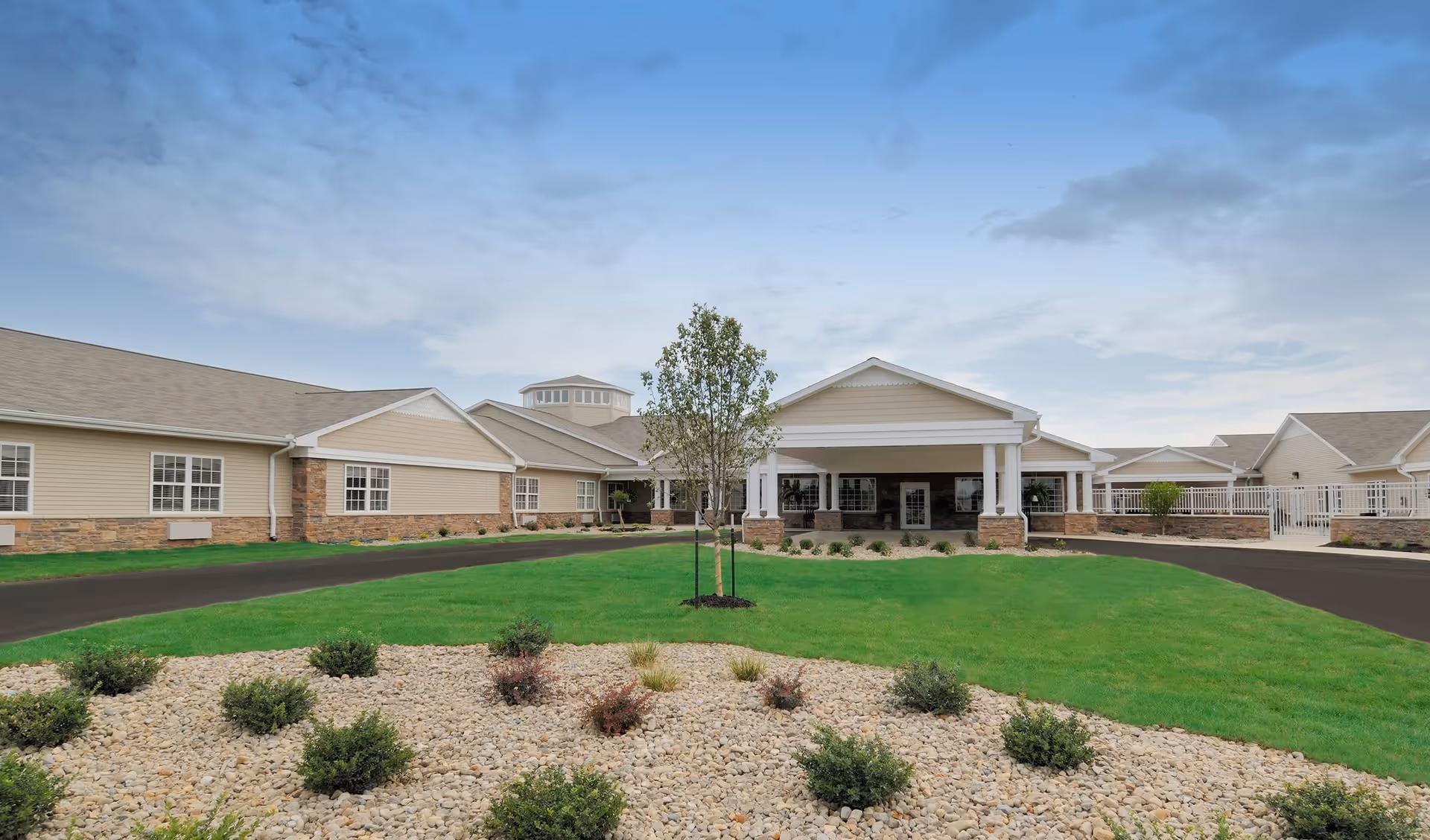 Exterior view of The Villages at Oak Ridge facility showing a single-story building with beige siding and stone accents, a covered entrance with white columns, a circular driveway, green lawn, and landscaped area with small bushes and rocks under a partly cloudy sky.