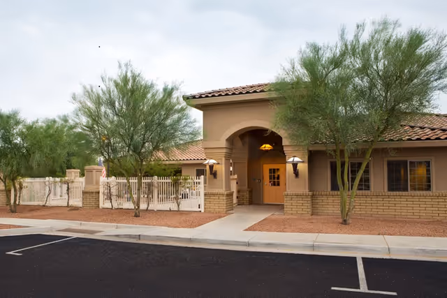 Exterior view of a beige senior living facility entrance with trees and a gate.