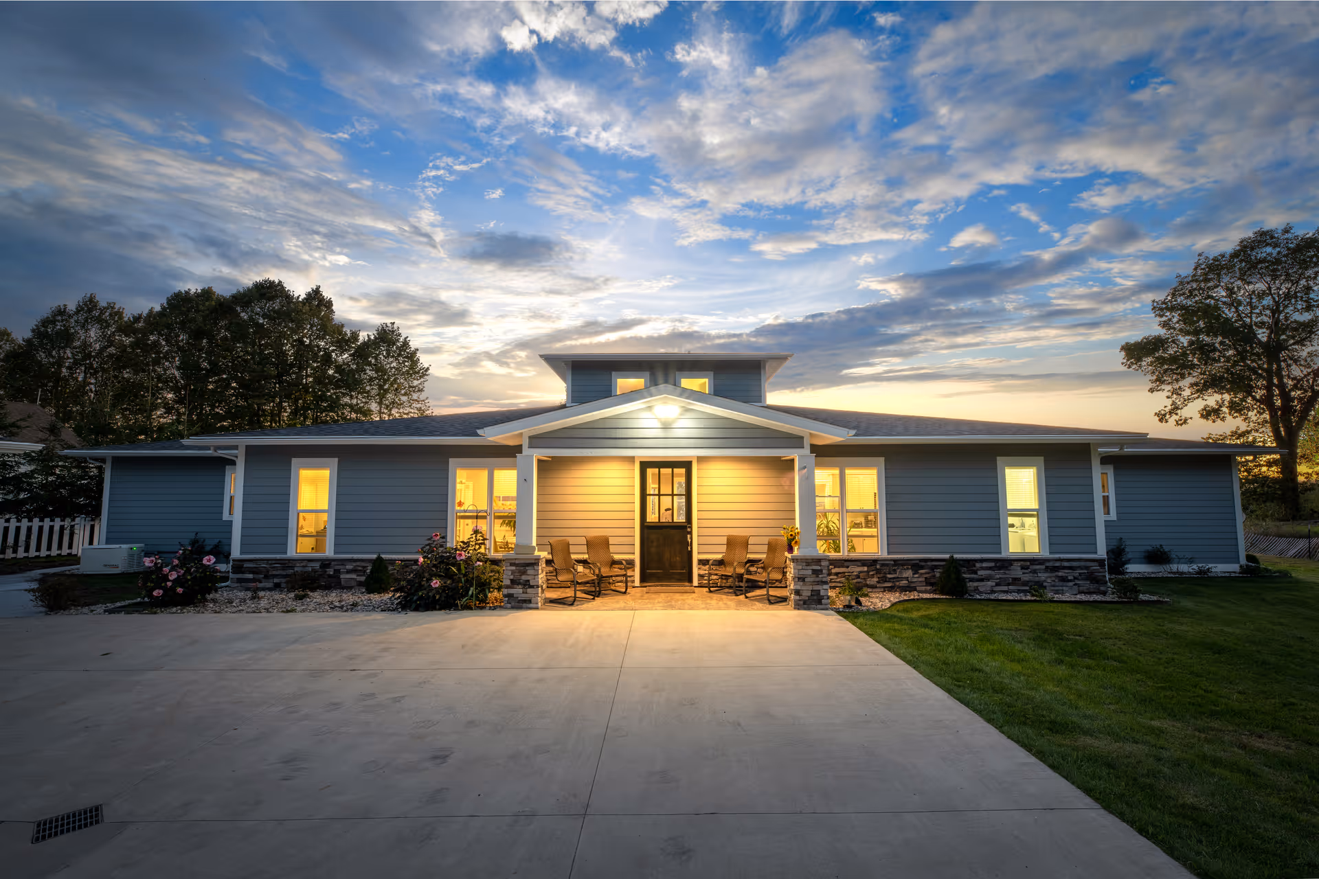 Exterior view of a single-story senior living facility building at dusk with lights glowing from inside. The building has a covered entrance with chairs on the porch, surrounded by a concrete driveway and green lawn, with trees in the background under a partly cloudy sky.