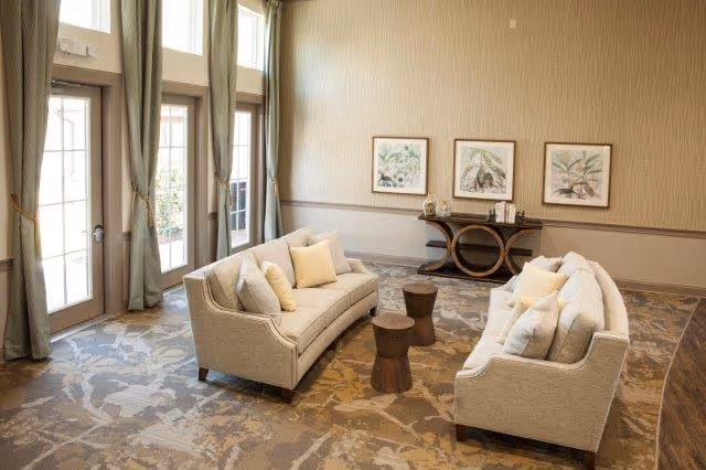 A bright and spacious living room area with two beige sofas facing each other, each adorned with multiple cushions. Between the sofas are two small wooden side tables. The room features large windows with green curtains allowing natural light to fill the space. On the far wall, there is a console table with decorative items and three framed botanical prints hanging above it. The floor is covered with a patterned carpet in neutral tones.