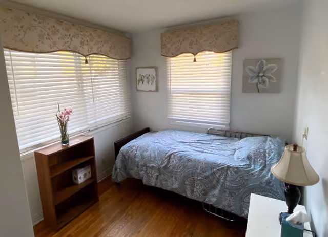 A small bedroom with a single bed, blue patterned bedspread, shelf, and large windows.