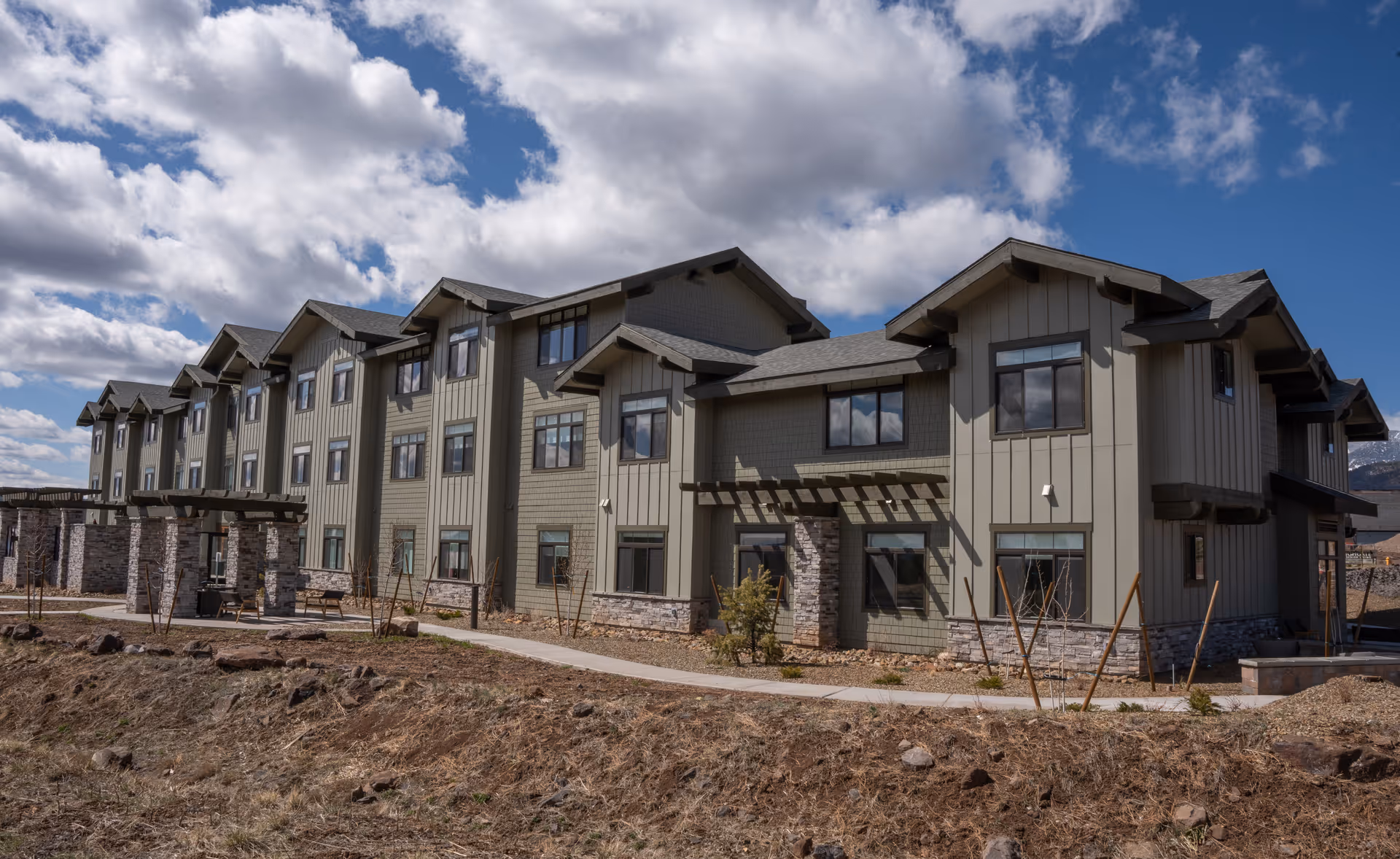 Exterior view of a multi-story senior living facility building with beige siding, stone accents, and multiple windows under a partly cloudy sky.