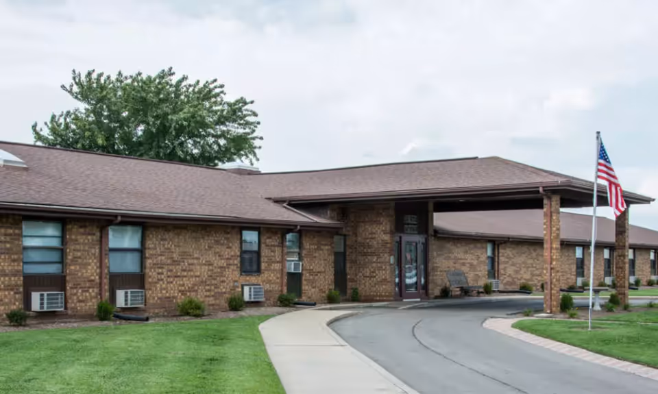 Exterior view of a single-story brick building with a covered entrance and an American flag on a flagpole in front. The building has several windows with air conditioning units and is surrounded by a well-maintained lawn and a sidewalk leading to the entrance.