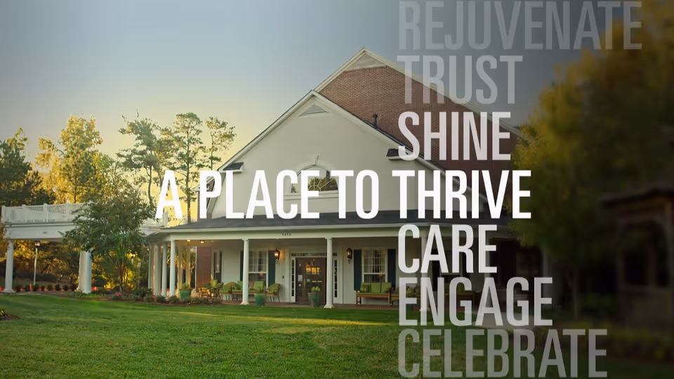Exterior view of a senior living facility building with a white facade, brick accents, a covered porch with columns, and outdoor seating. The building is surrounded by green grass and trees under a clear sky. Overlaid text reads: 'REJUVENATE TRUST SHINE A PLACE TO THRIVE CARE ENGAGE CELEBRATE'.