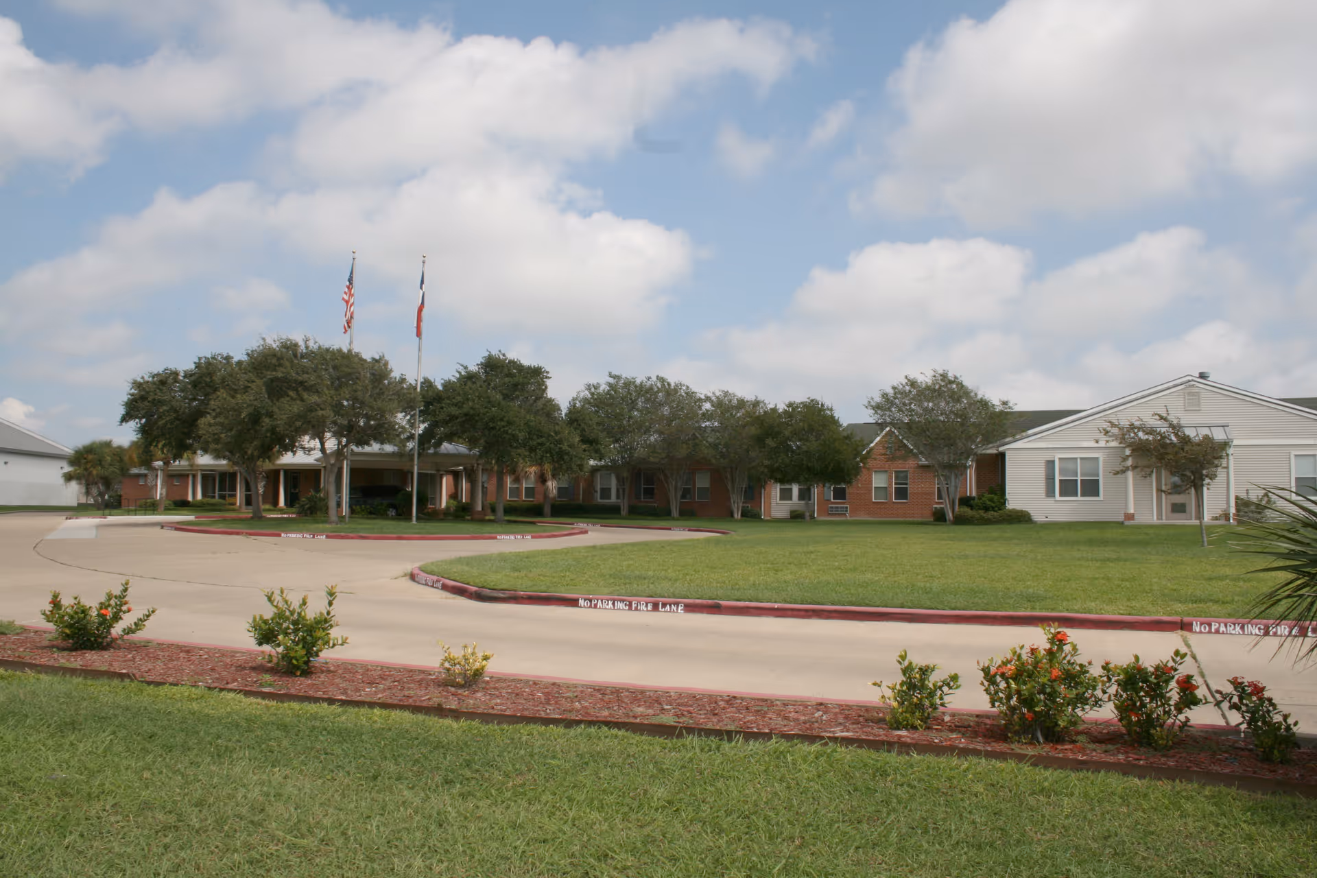 Exterior view of Villa of Corpus Christi South with flags, trees, and curved driveway.