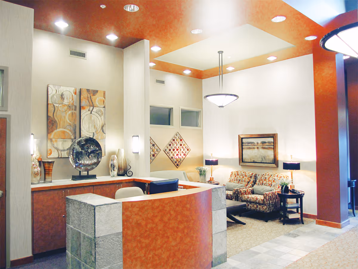 Reception area with a curved desk made of stone and wood, decorated with vases and art pieces on the wall. To the right, there is a seating area with two patterned armchairs, a small table with a lamp, and a framed landscape painting on the wall. The ceiling features recessed lighting and a hanging light fixture.